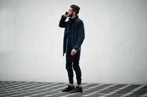 empresário do oriente médio usa casaco preto e camisa azul, óculos em pé contra a parede branca, fumando cigarro e fala por telefone. foto