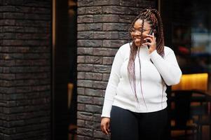 glamourosa mulher afro-americana na pose de suéter de gola alta branca na rua com telefone celular. foto