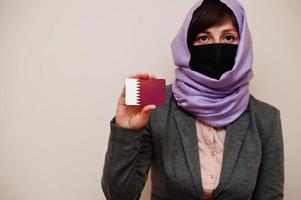 retrato de jovem muçulmana vestindo roupa formal, proteger máscara facial e lenço de cabeça hijab, segure o cartão de bandeira do catar contra fundo isolado. conceito de país coronavírus. foto