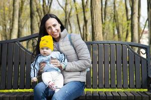 uma bela e jovem mãe atraente, com um menino, filho, está sentado em um banco em um parque de primavera. foto