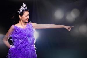 retrato do concurso de beleza do concurso de miss no vestido de noite de penas roxas com coroa de diamantes, mulher asiática aponta a mão do dedo para a direita com bokeh e fumaça sobre o espaço de cópia de fundo escuro foto