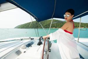 linda mulher de pele bronzeada de raça mista asiática anda ao longo de iates de luxo no oceano profundo, garota de óculos de sol de biquíni vermelho posando como modelo no cais de ancoragem sob o céu azul de verão nas férias foto