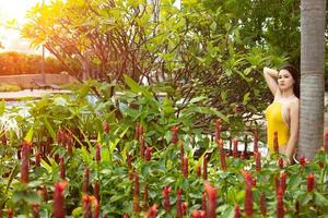 linda mulher sexy usar biquíni de maiô amarelo, mulher asiática relaxando no jardim do campo de flores vermelhas na piscina azul no hotel resort durante as férias de viagem, copie o espaço foto