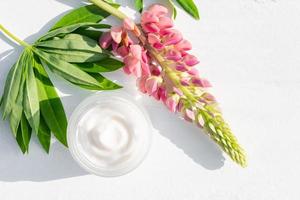 um frasco cosmético branco com creme natural de rosto em um fundo branco ao lado de tremoço rosa e folhas verdes. cosméticos orgânicos naturais para cuidados com a pele foto