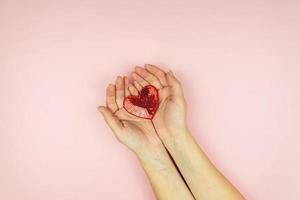 mãos femininas segurando um coração de lantejoulas vermelho sobre fundo rosa. layout mínimo criativo com espaço de cópia foto