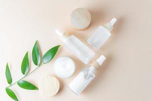 frasco de creme hidratante, soro em frasco de vidro e toner com folhas verdes sobre fundo bege. postura plana, vista superior. conjunto para produtos de beleza para cuidados com a pele e o corpo. pacote sem marca com rótulos em branco foto