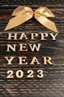 cartão de feliz ano novo 2023. letras de madeira e laço dourado sobre fundo escuro de madeira. foto