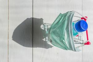 carrinho de compras em máscara médica com garrafa de desinfetante em fundo de madeira wgite. conceito de prevenção de coronavírus. vista do topo. foto