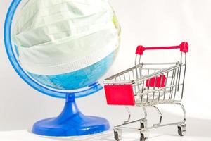 surto de coronavírus. pandemia mundial. globo em máscara de medicina e carrinho de compras em fundo branco. conceito de compras online durante o covid-19. foto