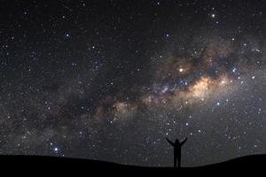paisagem com Via Láctea, céu noturno com estrelas e silhueta de um homem desportivo em pé com os braços levantados na montanha alta.
