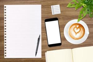 um caderno em branco aberto, smartphone com caneta e uma xícara de café com leite na mesa de madeira.