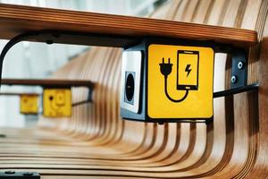 um banco de madeira de designer com tomadas e uma placa amarela para carregar smartphones na sala de espera. foto