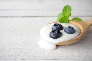 iogurte branco com folha de hortelã e mirtilo fresco na colher de pau no fundo branco de madeira.