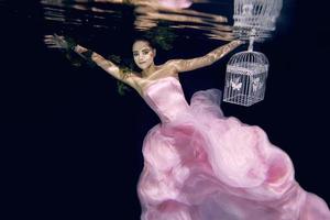 jovem mulher bonita de vestido longo rosa com gaiola nas mãos debaixo d'água foto