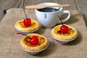 torta de ovo em copo de papel alumínio com cereja vermelha e café na mesa de madeira foto