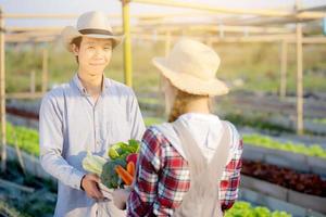 bela jovem asiática e homem segurando vegetais orgânicos frescos com cesta juntos na fazenda hidropônica, colheita e agricultura e cultivo para alimentação saudável e conceito de negócios. foto