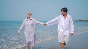 casal de aposentados asiáticos relaxantes à beira-mar no verão. uma mulher sênior andando e de mãos dadas com seu parceiro na praia. foto