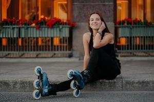 tiro horizontal de jovem ativo faz uma pausa enquanto anda de rolos na rua vestida com roupas esportivas pretas sente sorrisos satisfeitos agradavelmente poses ao ar livre. estilo de vida de verão e conceito de descanso foto