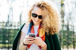 mulher bonita com cabelos cacheados, usando óculos escuros e jaqueta verde, segurando o telefone inteligente nas mãos, navegando na internet, desfrutando de comunicação on-line em pé contra o fundo do parque verde foto