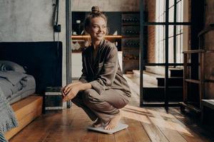 imagem do alegre modelo satisfeito jovem em pijama aconchegante de cetim agachado sobre balanças com um largo sorriso no quarto moderno foto
