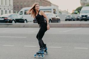 tiro lateral de jovem ativa em patins de boa forma física ao longo das ruas da cidade parece feliz por trás usa roupas esportivas pretas leva um estilo de vida saudável. conceito de recreação e hobby foto