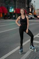 satisfeito mulher europeia mantém a mão na cintura sorrisos agradavelmente leva estilo de vida ativo passeios de patins vestido com roupas pretas poses contra fundo desfocado da cidade movimentada. conceito de esporte radical foto