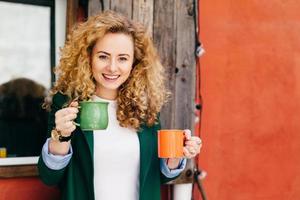 mulher feliz com cabelos loiros cacheados fofos encantadores olhos azuis segurando duas canecas de café vai tratar seus amigos com café tendo expressão satisfeita e sorriso no rosto. conceito de linguagem corporal foto