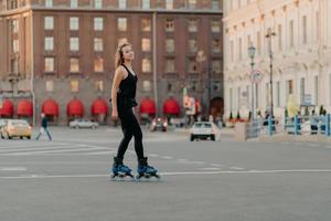 tiro de corpo inteiro de patins de mulher de cabelos escuros magros na estrada de asfalto vestida em roupas esportivas pretas gosta de exercícios de atividades ao ar livre para um corpo saudável e forte. conceito de esporte e hobby foto