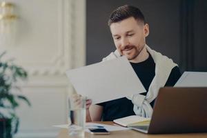 tiro interno de freelancer masculino concentrado em papéis prepara projeto ou relatório financeiro posa no desktop com computador portátil trabalha em casa fundo desfocado aconchegante usa roupas casuais. foto