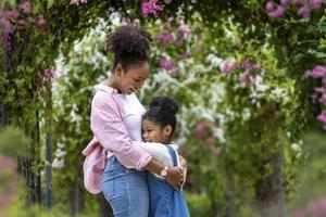 mãe e filha africanas se abraçando enquanto caminhavam alegremente sob o arco de treliça de flores no parque público durante o verão para o amor e cuidado da família na celebração do dia das mães foto