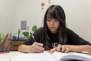 aluna asiática está escrevendo lição de casa e lendo livro na mesa foto