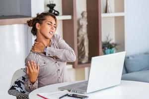 retrato de uma jovem estressada sentada na mesa de escritório em casa na frente do laptop, tocando as costas doloridas com expressão de dor, sofrendo de dor nas costas depois de trabalhar no pc foto