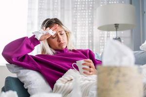 mulher.flu.mulher doente pegou resfriado. espirrar no tecido. dor de cabeça. vírus .medicina. jovem infectada com frio assoando o nariz no lenço. mulher doente com dor de cabeça sentada em um sofá foto