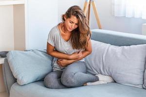 mulher deitada no sofá parecendo doente na sala de estar. bela jovem deitada na cama e de mãos dadas na barriga dela. mulher com dor de estômago dolorosa na cama, período menstrual foto