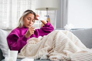 doença, conceito de problema de vírus sazonal. mulher doente com gripe deitada no sofá olhando a temperatura no termômetro. mulher doente deitada na cama com febre alta. gripe fria e enxaqueca. foto