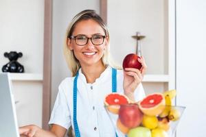 retrato de jovem nutricionista feminina sorridente na sala de consulta. mesa de nutricionista com frutas saudáveis, suco e fita métrica. nutricionista trabalhando no plano de dieta. foto