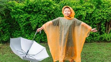 menino asiático vestindo capa de chuva laranja segurando guarda-chuva feliz e se divertindo na chuva em um dia chuvoso foto