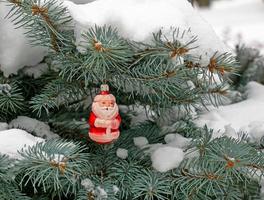 brinquedo de natal papai noel pendura em um galho coberto de neve de uma árvore de natal em um fundo festivo de neve branca bokeh com espaço de cópia. foto