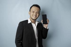 um retrato de um empresário asiático feliz está sorrindo e segurando seu smartphone mostrando espaço de cópia na tela vestindo terno preto isolado por um fundo azul foto