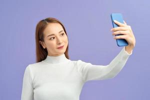 mulher jovem e atraente em roupas casuais fazendo selfie por seu telefone inteligente e sorrindo em pé contra o fundo violeta foto