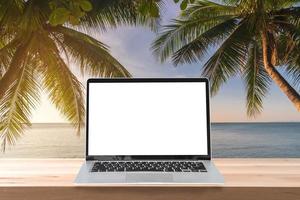 laptop de tela vazia em uma mesa de madeira com coqueiro e pôr do sol na praia tropical, conceito de férias de verão foto