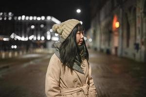 uma jovem e bela mulher coreana elegante de chapéu e casaco caminha pelas ruas da cidade à noite. foto