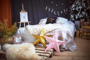 decoração de interiores de natal em estilo loft. uma cama sobre um estrado, uma grande manta tricotada, estrelas douradas e cor-de-rosa, guirlandas, um cavalete. árvore de natal com caixas de presente no quarto. clima de ano novo foto