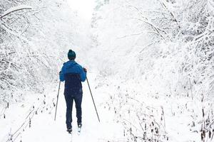 esquiador de blusão e chapéu com pompom com bastões de esqui nas mãos com as costas no contexto de uma floresta de neve. esqui cross-country na floresta de inverno, esportes ao ar livre, estilo de vida saudável. foto