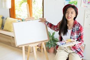 jovem sentada em uma cadeira com cavalete para desenhar segure a paleta de cores e pincel na sala. foto