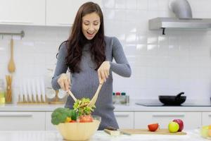 mulher bonita, legal e grávida está felizmente cozinhando saladas orgânicas e saudáveis na cozinha moderna. conceito de cuidados com o corpo e preparação antes do parto foto