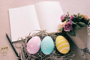 ovos de páscoa pastel e coloridos no ninho e buquê de flores com caderno em branco sobre fundo de madeira, feliz conceito de férias de páscoa foto