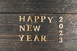feliz ano novo 2023 celebração. texto de madeira em fundo de madeira com confetes dourados espalhados foto