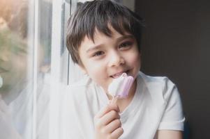 uma criança feliz comendo um sorvete, retrato de um menino bonito sentado ao lado de uma janela tomando um refresco, criança com um sorriso no rosto relaxando em casa. criança olhando para a câmera foto