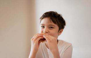 criança saudável comendo sanduíches de bacon caseiro com legumes misturados, menino feliz tomando café da manhã em casa antes de ir para a escola foto